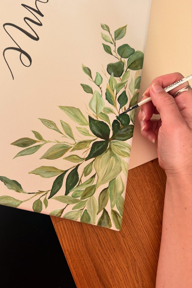 Kirsty hand-painting eucalyptus foliage onto a cream canvas welcome sign
